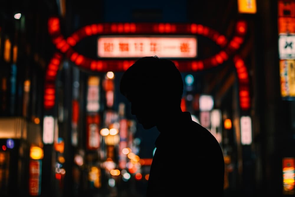夜の新宿歌伎町のネオンがぼんやりと光り、その手前に立つ人物のシルエットが浮かび上がるアーティスティックな写真
